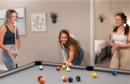 One young women takes a shot at pool while two other women watch